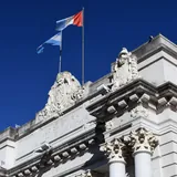 Detalle superior de la fachada de la Legislatura de la Provincia de Santa Fe, destacando sus columnas, esculturas ornamentales y las banderas de Argentina y de Santa Fe flameando bajo un cielo azul.