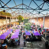 Vista aérea de una feria con puestos techados de violeta y banderines de colores dentro de la estructura abierta del Mercado Progreso.