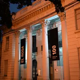 Fachada iluminada de noche del Museo Provincial de Bellas Artes Rosa Galisteo de Rodríguez en Santa Fe, destacando sus grandes columnas clásicas con carteles negros verticales.