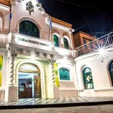 Vista nocturna de la fachada iluminada del Club de Regatas Santa Fe, con sus paredes blancas, columnas salomónicas en la entrada y ventanas de arco verde.